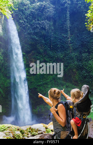 Young happy woman hold little traveller in back baby carrier, explore jungle waterfall in rainforest. Stock Photo