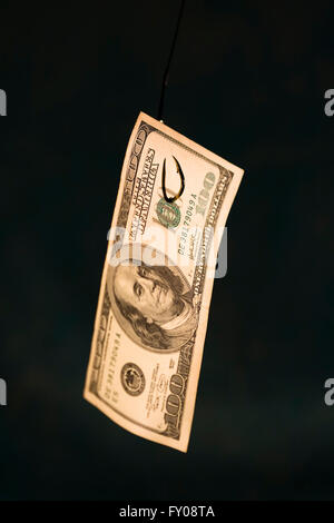 A 100 dollar bill hanging on a fishing hook sharp focus on hook coming through paper on dark blue background Stock Photo