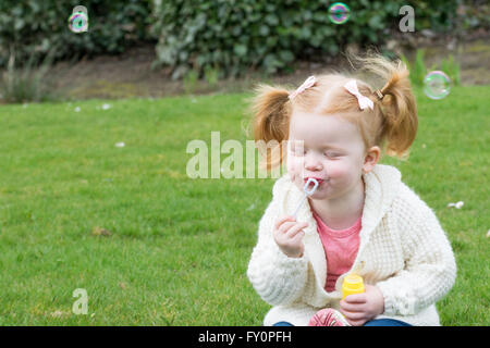 toddler girl blowing bubbles while sitting on grass Stock Photo