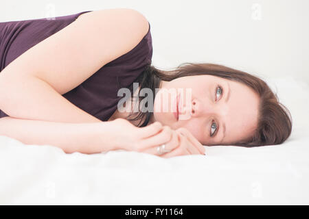Portrait of young woman lying down in her bed resting. She is looking away with a serious and pensive expression. Stock Photo