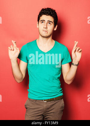 Young man crossing fingers while looking up against red background Stock Photo