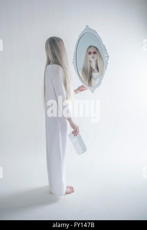 Young woman with spooky red eyes looking in mirror against white background Stock Photo