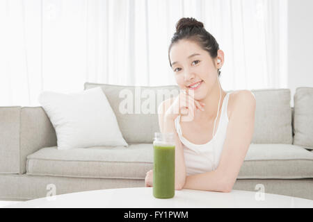 Portrait of young smiling woman posing with green vegetable juice against sofa and curtains Stock Photo