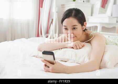 Young smiling woman listening to music on smartphone in bed Stock Photo