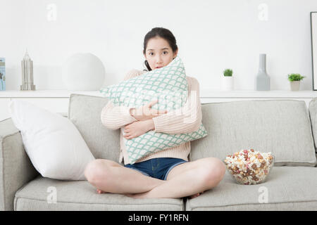 Young woman sitting on sofa hugging a cushion with frightened face Stock Photo