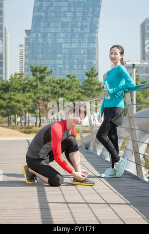 Side view of young smiling man tying his shoelaces staring at front and young smiling woman standing staring at front Stock Photo