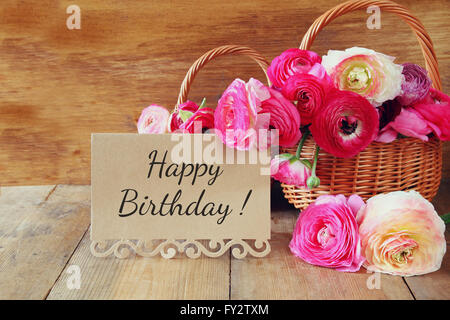 pink flowers in the basket next to card with phrase: happy birthday, on wooden table.