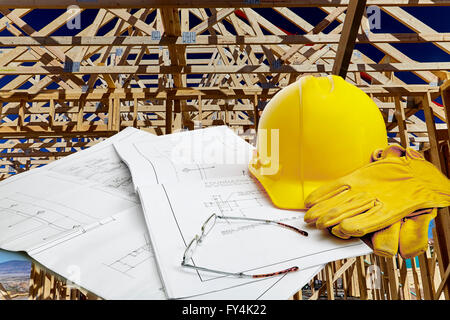 A set of blueprints with hardhat, work gloves and reading glasses with wood framed structure in background Stock Photo