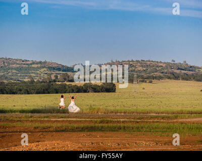 A bride and her bridesmaid dressed in their wedding attire jump for joy at sunset in a large open field in Chico, California. Stock Photo