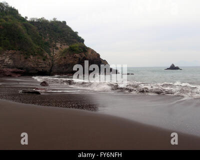 Waves crash on empty dark sand beach in Manzanillo, Mexico with a rock island and rocky cliff surrounding. Stock Photo