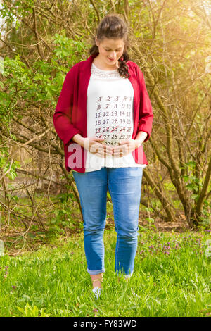 Pregnant woman in red jacket with calendar on her T-shirt outdoor in the park. Stock Photo