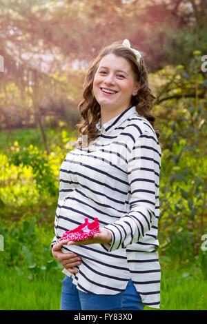 Close up of a pregnant woman holding baby shoes in her hands in a park Stock Photo