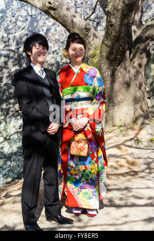 Japanese young married couple. Man in formal western style suit, woman in red kimono. Standing under cherry blossom tree, smiling. No eye-contact. Stock Photo