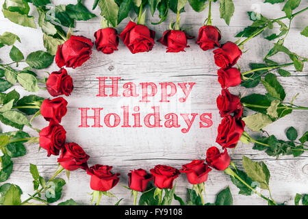 Happy holidays text in circle made of red roses on white rustic wooden table. Top view. Stock Photo