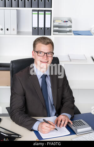 Young businessman is writing into a file while sitting at the desk in the office. The man is looking to the camera. Stock Photo