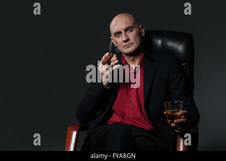 Serious handsome businessman drinking whiskey and smoking cigar sitting in office chair over black background Stock Photo