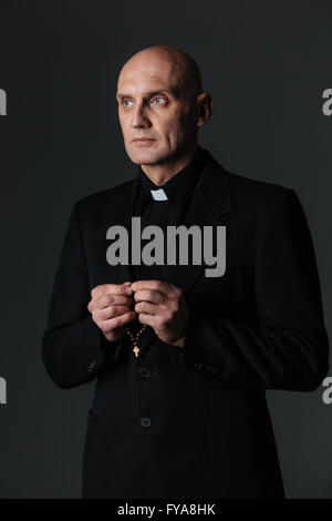 Portrait of handsome priest standing and holding rosary over black background Stock Photo