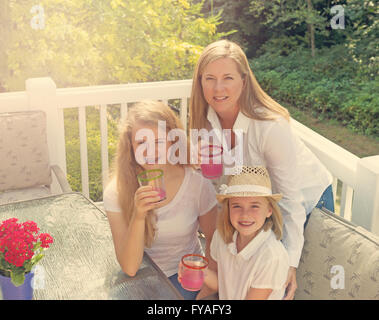 Top front view of happy mother and daughters, looking forward, drinking grapefruit juice while outdoors on patio with woods. Stock Photo