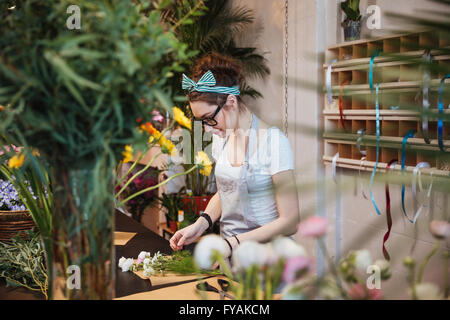 Beautiful young woman florist in apron and glasses making bouquet and working in flower shop Stock Photo