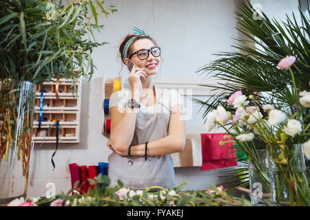 Cheerful cute young woman florist standing and talking on mobile phone in flower shop Stock Photo