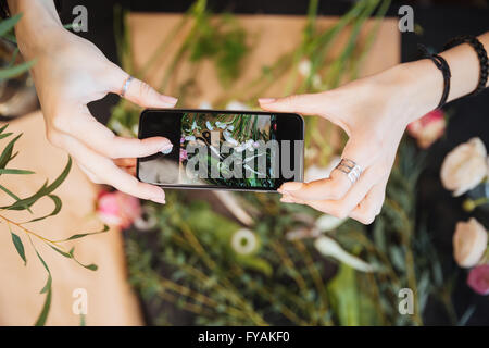 Top view of hands of young woman florist holding smartphone and taking photos of flower on table