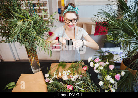 Happy beautiful young woman taking photos of flowers on table in the shop Stock Photo