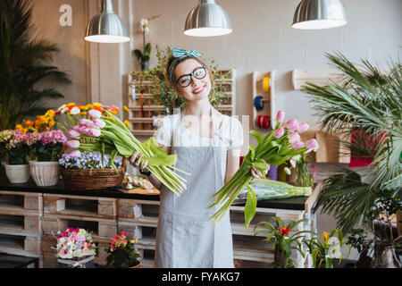 Cheerful charming young woman florist standing and holding two bunches of pink tulips in flower shop Stock Photo