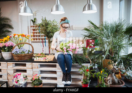 Cute lovely young woman florist sitting and arranging pink tulips in flower shop Stock Photo