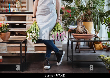 Closeup of young woman florist in apron and sneakers with two bouquets of tulips standing in flower shop Stock Photo