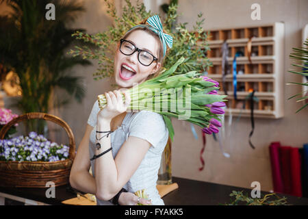 Playful happy young woman florist holding bunch of purple tulips and winking in flower shop Stock Photo