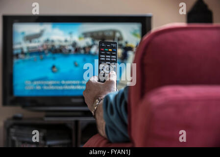 Man in comfy chair switching channels with remote control to look for pleasing programme on television set in living room Stock Photo