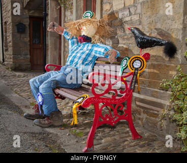 Scarecrow sat on a red bench.  Best Crow, Bench & Hat Wray's Annual Scarecrow Festival.  The villagers are back again having created some weird, wacky and wonderful scarecrow creations to  delight visitors. Annually Wray village hosts a display of scarecrows to fit in with a subject on the school curriculum, with creations popping up on village gardens, peeking out of windows and perching on balconies. The Scarecrow Festival, established 1995, takes place during the week leading up to May Day when there is a fair and a weekend parade. Stock Photo