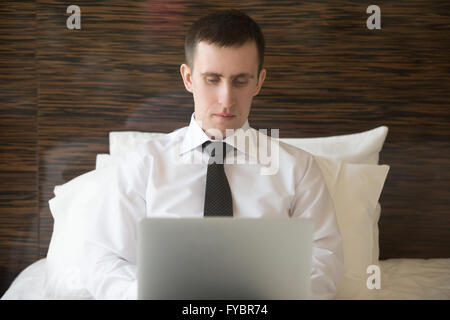 Portrait of serious handsome young business man sitting on pillow on bed and working at home or in trip on his laptop computer Stock Photo
