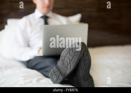 Handsome young business man sitting on bed in formal wear and his shoes on. Businessman working at home or in trip on his laptop Stock Photo