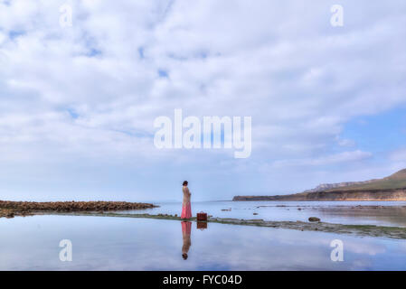 a woman in a vintage pink dress is standing at the sea with her suitcase Stock Photo