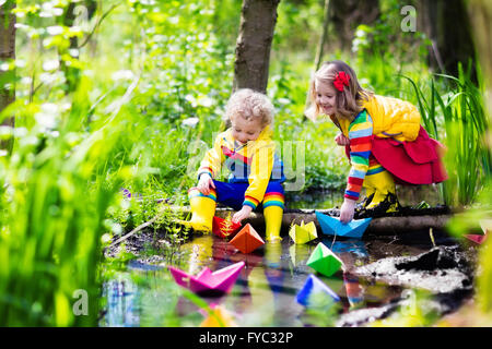 Children play with colorful paper boats in a small river on a sunny spring day. Kids playing exploring the nature. Stock Photo