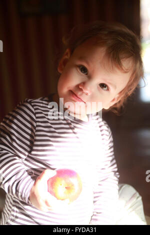a 15 months young kid enjoys time with an apple in hand sitting in the rays of spring sun Stock Photo
