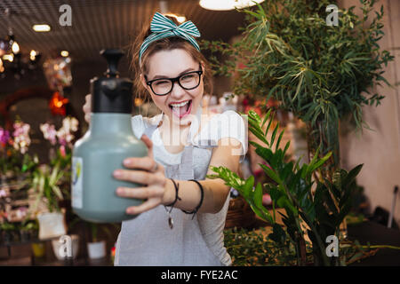 Playful attractive young woman florist pointing with water sprayer on you in flower shop Stock Photo