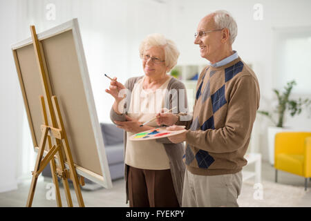 Senior couple drawing together on a canvas and looking at the painting at home Stock Photo