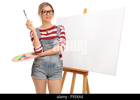 Young blond woman holding a paintbrush in front of a blank canvas isolated on white background Stock Photo