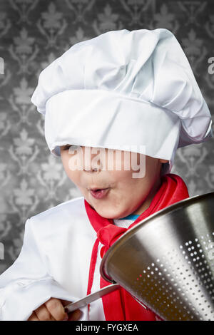 Funny boy cook in uniform over vintage  background playing with cookware Stock Photo