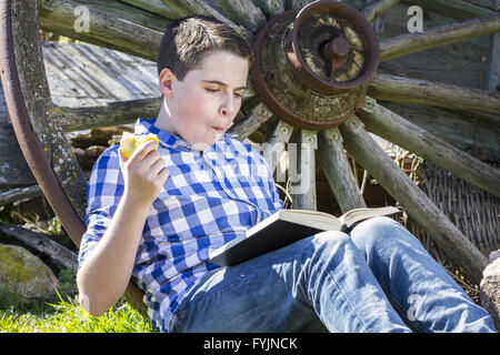 Joyful.Young boy reading a book in the woods eating an apple Stock Photo