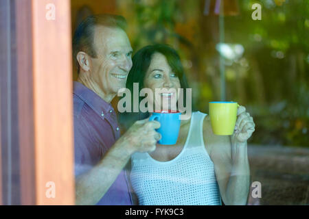 Senior man and woman looking out of the tinted glass doors of their home while drinking coffee from mugs Stock Photo