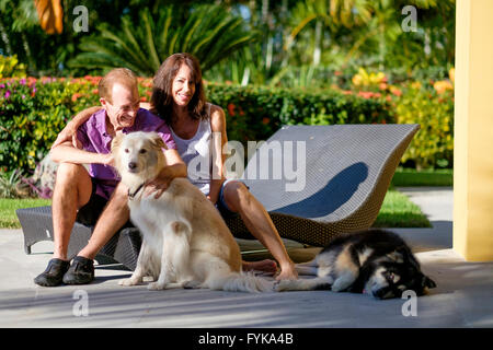 Retired senior man and woman petting their 2 dogs while sitting on lounge chairs in the backyard of their home in Mexico Stock Photo