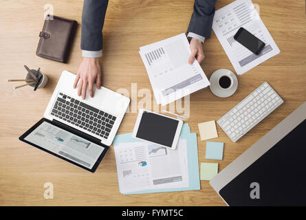 Businessman working at office desk and checking financial reports with computers and paperwork around, top view Stock Photo
