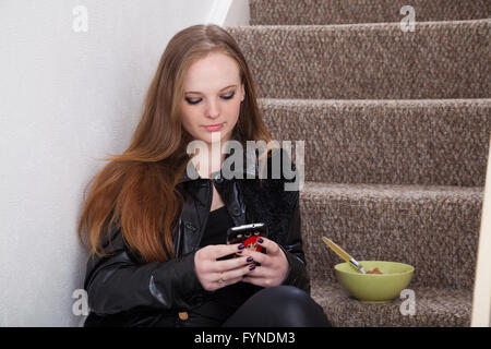 A long haired redheaded woman sitting on the stairs of her house using her mobile phone with a bowl of cereal next to her. Stock Photo