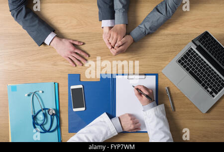 Doctor writing down medical records and talking with a couple during a visit, desktop top view, unrecognizable people Stock Photo