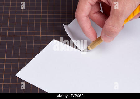 A hand holding a utility knife with a yellow handle cutting a piece of white paper on a cutting mat. Stock Photo