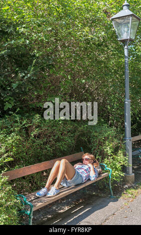 Middle aged caucasian tanned sad woman in sun glasses lying outdoor on park bench. Stock Photo