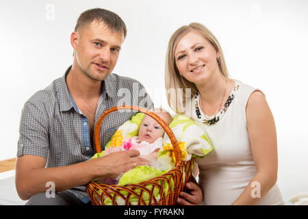 Mom and dad sit on the bed and holding a two-month baby girl in a basket Stock Photo